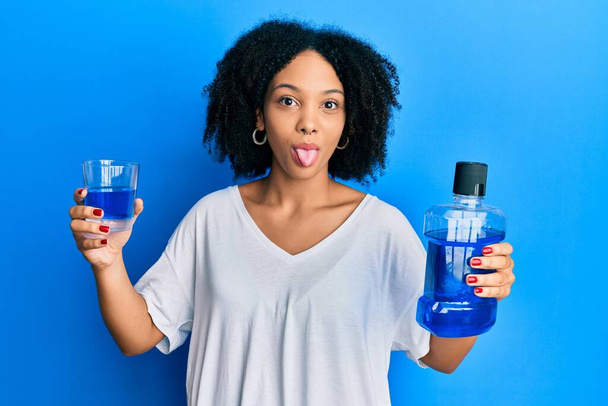 Young african american girl holding mouthwash for fresh breath sticking tongue out happy with funny expression.  - Photo, Image