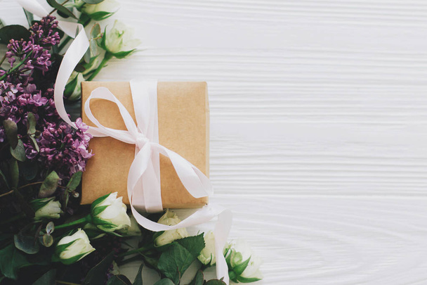 Κομψό κουτί δώρου με λιλά και τριαντάφυλλα μπουκέτο επίπεδη θέσει σε λευκό ξύλινο φόντο, χώρο για κείμενο. Ευτυχισμένη ημέρα των μητέρων και έννοια ημέρα των γυναικών. Χειροτεχνία με κορδέλα και ανοιξιάτικα λουλούδια - Φωτογραφία, εικόνα
