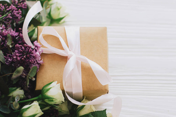 Счастливых матерей и женщин. Стильная подарочная коробка с сиренью и букет роз на белом деревянном фоне в мягком свете, пространство для текста. Ремесло подарено лентой и весенними цветами - Фото, изображение