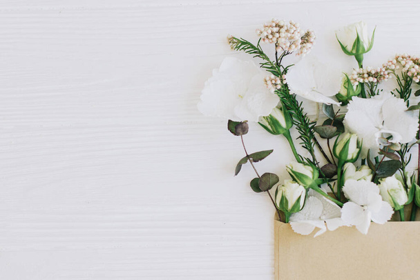 Stijlvolle rozen, hortensia en eucalyptus takken groeien van ambachtelijke envelop op wit hout, plat lag met kopieerruimte. Creatief voorjaarsbeeld, bloemenwenskaart en bloemlevering - Foto, afbeelding