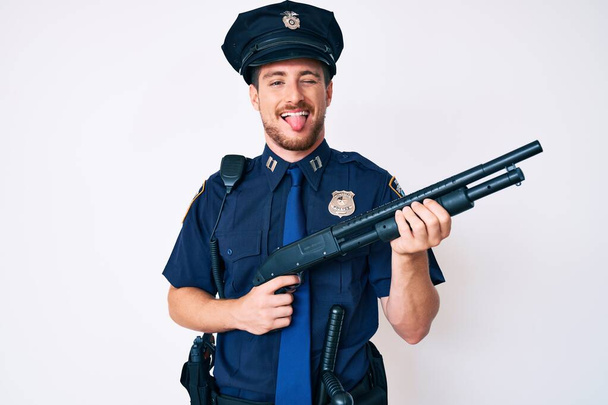 Jeune homme caucasien portant uniforme de police tenant fusil de chasse collant la langue heureux avec une expression drôle.  - Photo, image