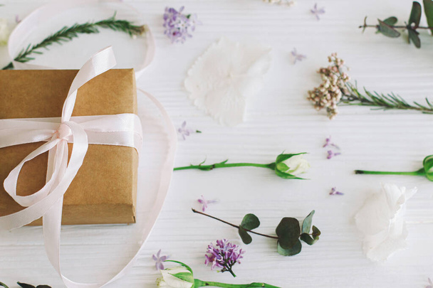 Happy mothers day and womens day concept. Craft present with ribbon and modern floral composition. Stylish gift box with lilac and roses flowers and eucalyptus leaves  on white wood - 写真・画像