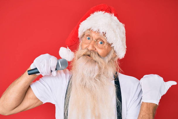 Alter älterer Mann mit grauen Haaren und langem Bart im Weihnachtsmann-Kostüm singt, das Mikrofon zeigt den Daumen zur Seite und lächelt glücklich mit offenem Mund.  - Foto, Bild