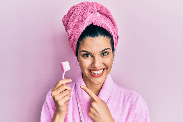 Fiatal spanyol nő zuhanyköntöst visel, fogkefét tart, mosolyogva, boldogan mutatva kézre és ujjra.  - Fotó, kép