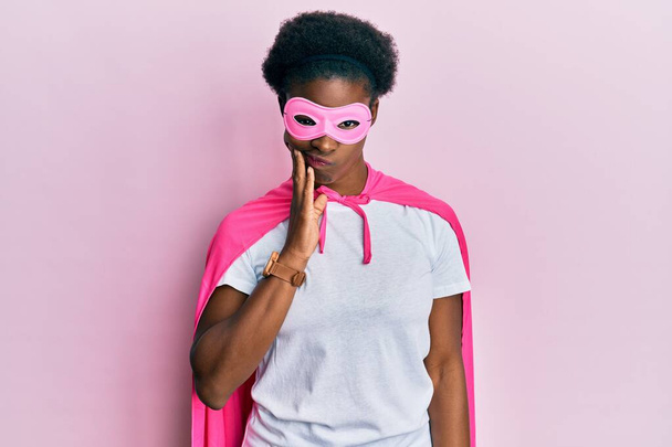 歯の痛みや歯の病気のために痛みを伴う表現で手で口に触れるスーパーヒーローマスクとケープ衣装を身に着けている若いアフリカ系アメリカ人の女の子.歯科医  - 写真・画像