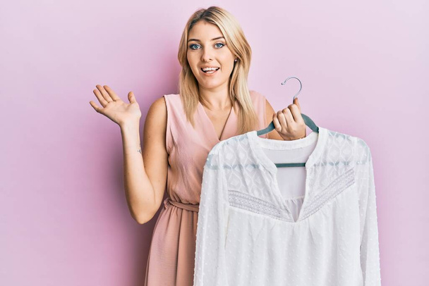 Young caucasian woman holding hanger with t shirt celebrating achievement with happy smile and winner expression with raised hand  - Фото, изображение
