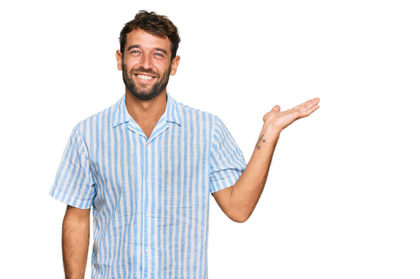 Knappe jongeman met baard in casual fris shirt lachend vrolijke presentatie en wijzend met handpalm kijkend naar de camera.  - Foto, afbeelding