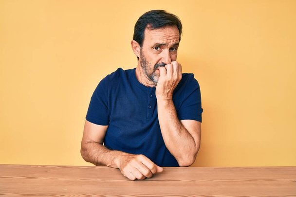 Középkorú spanyol férfi alkalmi ruhát visel az asztalon ülve, stresszesnek és idegesnek tűnik, kezével a száján körmöket harap. szorongás.  - Fotó, kép