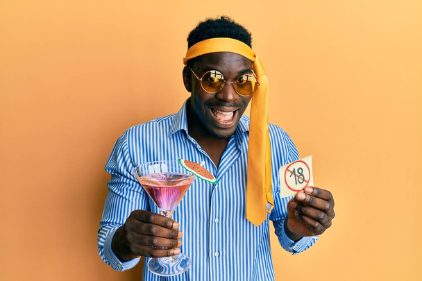 Handsome black man drunk wearing tie over head drinking cocktail holding under 18 sign celebrating crazy and amazed for success with open eyes screaming excited.  - Zdjęcie, obraz