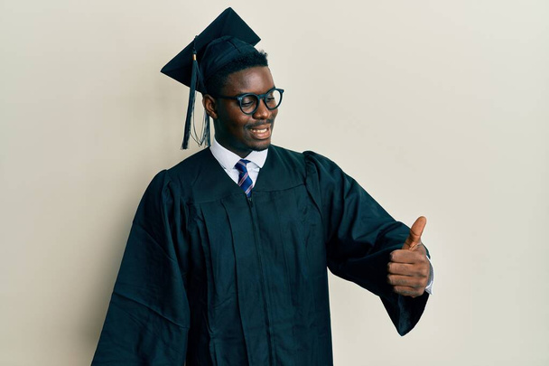 Handsome black man wearing graduation cap and ceremony robe looking proud, smiling doing thumbs up gesture to the side  - Foto, immagini