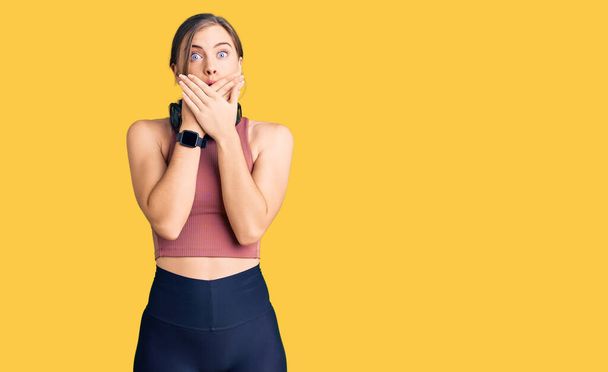 Beautiful caucasian young woman wearing gym clothes and using headphones shocked covering mouth with hands for mistake. secret concept.  - Fotografie, Obrázek