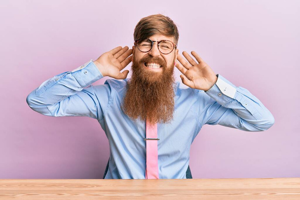 Молодой ирландский рыжий мужчина в деловой рубашке и галстуке сидит на столе, пытаясь услышать обе руки на ушной жест, любопытный для сплетен. проблема со слухом, глухой  - Фото, изображение