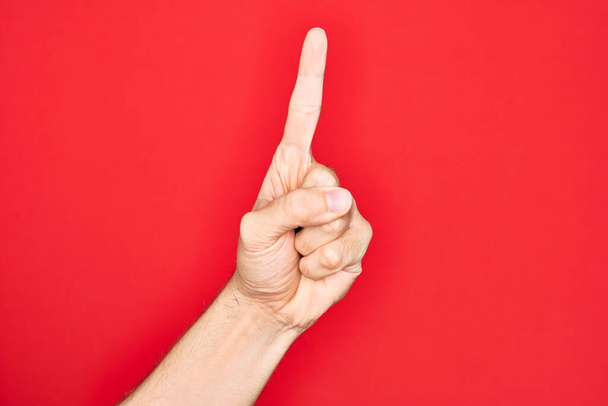 Hand van Kaukasische jongeman met vingers over geïsoleerde rode achtergrond tellend nummer één met behulp van wijsvinger, met idee en begrip - Foto, afbeelding