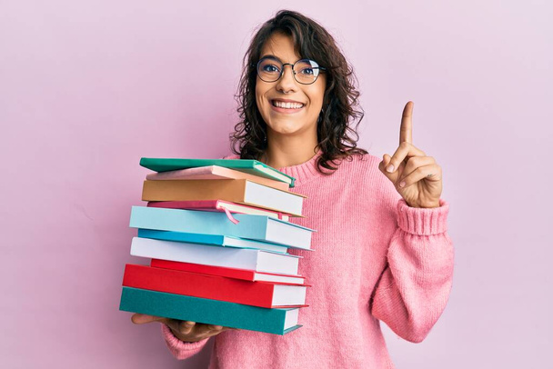 Junge hispanische Frau mit einem Stapel Bücher, die mit einer Idee oder Frage lächelt und mit glücklichem Gesicht den Finger zeigt, Nummer eins  - Foto, Bild