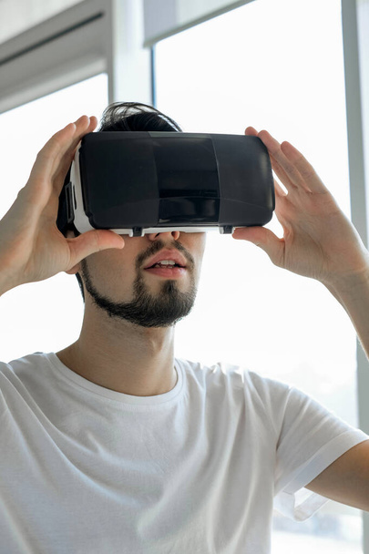 Un giovane gioca vigorosamente a un videogioco di realtà virtuale indossando occhiali VR. Quell'uomo gioca ai videogiochi. Rilassati. Fine settimana pieno di gioia. Gioco multigiocatore. Giochi online. Streaming di videogiochi. - Foto, immagini