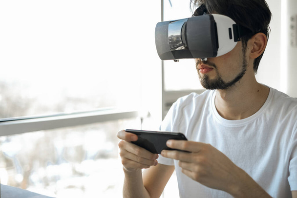 Young man vigorously plays virtual reality video game wearing VR glasses at home. Multiplayer game. Online Games. Streaming video games. The man plays video games. Time relax. Joyfull weekend.  - Photo, Image