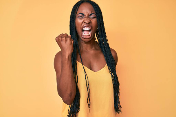African american woman with braids wearing casual clothes angry and mad raising fist frustrated and furious while shouting with anger. rage and aggressive concept.  - Foto, afbeelding