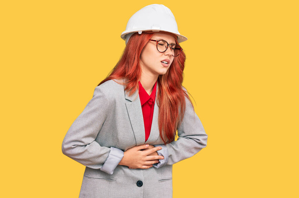 Nuori punapää nainen yllään arkkitehti kypärä käsi vatsaan, koska pahoinvointi, kivulias sairaus huonovointisuus. särky käsite.  - Valokuva, kuva