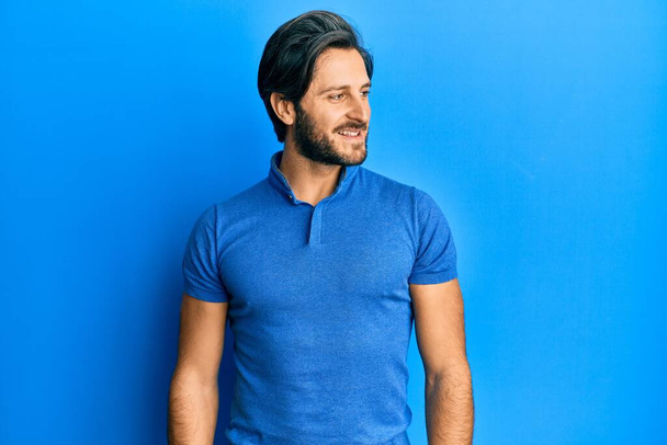 Latin kökenli genç bir adam günlük mavi tişörtler giyiyor. Yan tarafa bakıyor, rahat bir görünüm takınıyor ve kendine güvenen bir gülümseme takınıyor..  - Fotoğraf, Görsel