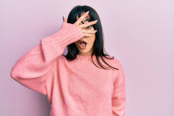 Young brunette woman with bangs wearing casual winter sweater peeking in shock covering face and eyes with hand, looking through fingers with embarrassed expression.  - Фото, изображение