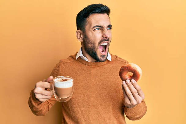 Jonge Spaanse man die donut eet en koffie drinkt boos en gek schreeuwend gefrustreerd en woedend, schreeuwend van woede. woede en agressief concept.  - Foto, afbeelding