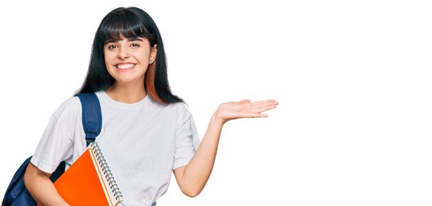 Menina hispânica jovem usando mochila estudantil e segurando livro celebrando a vitória com sorriso feliz e expressão vencedora com as mãos levantadas  - Foto, Imagem