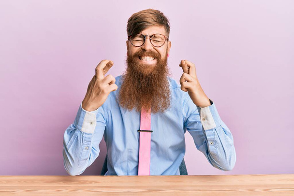 Junger irischer Rotschopf in Businesshemd und Krawatte sitzt auf dem Tisch und gestikuliert mit erhobenem Zeigefinger lächelnd mit geschlossenen Augen. Glück und abergläubisches Konzept.  - Foto, Bild