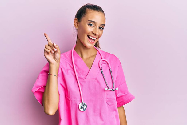 Mujer hispana joven vistiendo uniforme médico y estetoscopio mostrando y señalando con el dedo número uno mientras sonríe confiada y feliz.  - Foto, Imagen
