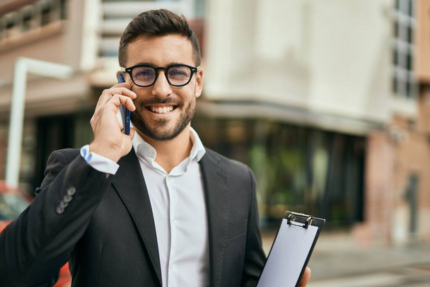 Молодой латиноамериканец-бизнесмен улыбается счастливо разговаривая на смартфоне в городе. - Фото, изображение
