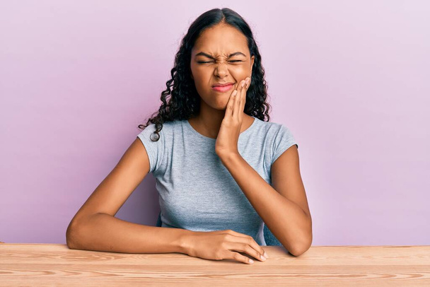 Jeune fille afro-américaine portant des vêtements décontractés assis sur la table touchant la bouche avec une expression douloureuse à cause de maux de dents ou de maladies dentaires sur les dents. dentiste  - Photo, image