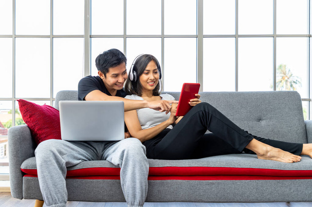Young Asian couple relaxing sitting on sofa using laptop computer and digital tablet working from home - Foto, afbeelding