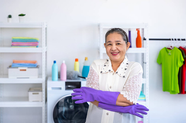 Asiatische Seniorin mit Gummihandschuhen und Armbrust in Waschküche mit stolzem Blick in die Kamera, Frühjahrsputz-Konzept - Foto, Bild