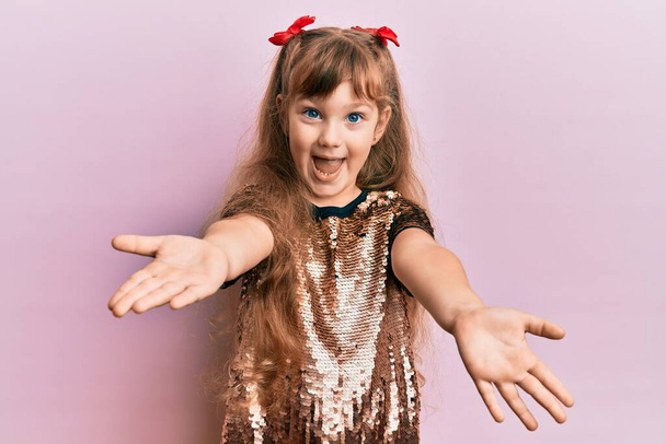 Kis kaukázusi lány gyerek ünnepi flitteres ruhában nézi a kamerát, ahogy tárt karokkal mosolyog. vidám kifejezés, mely felöleli a boldogságot.  - Fotó, kép
