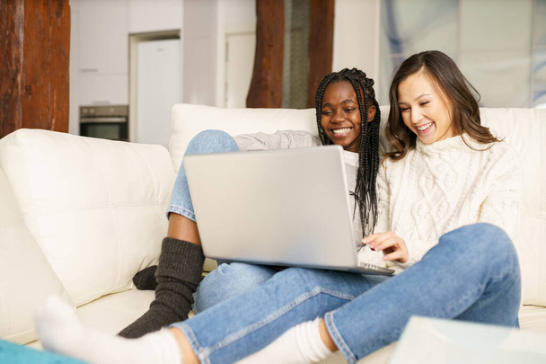 Two female student friends sitting on the couch at home using a laptop. - Photo, image