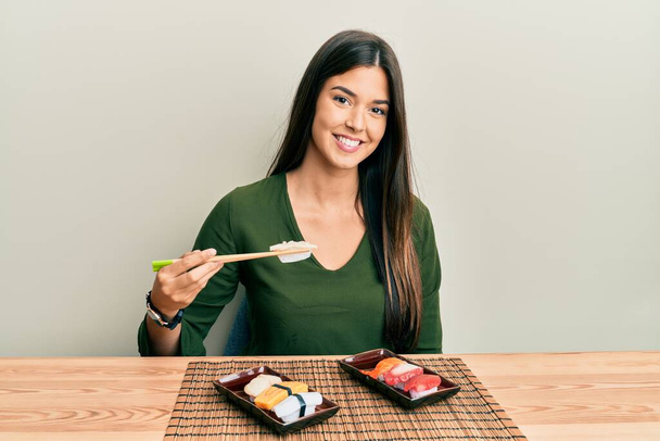 Young brunette woman eating sushi sitting on the table looking positive and happy standing and smiling with a confident smile showing teeth  - Foto, afbeelding