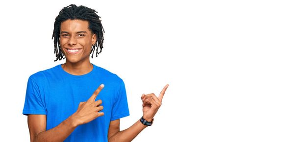 Junger afrikanisch-amerikanischer Mann in lässiger Kleidung lächelt und blickt mit zwei Händen und Fingern zur Seite in die Kamera.  - Foto, Bild
