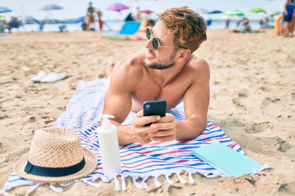 Handsome fitness caucasian man at the beach on a sunny day sunbathing lying on the towel using smartphone - Zdjęcie, obraz