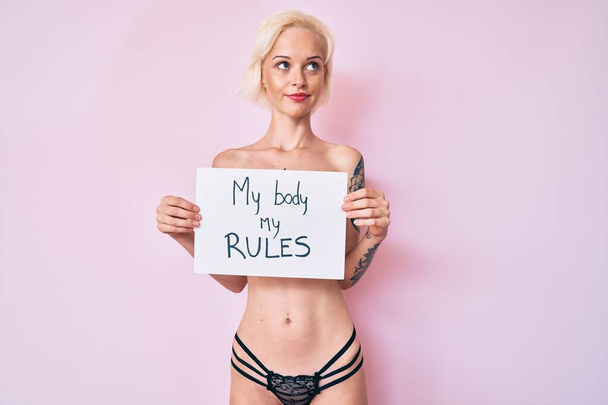 Young blonde woman with tattoo standing shirtless holding banner with my body my rules message smiling looking to the side and staring away thinking.  - Foto, afbeelding