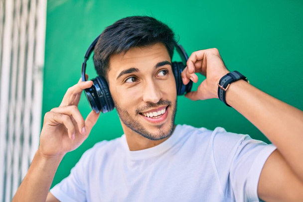 Młody Latynos uśmiecha się szczęśliwy używając słuchawek opierających się na ścianie w mieście. - Zdjęcie, obraz