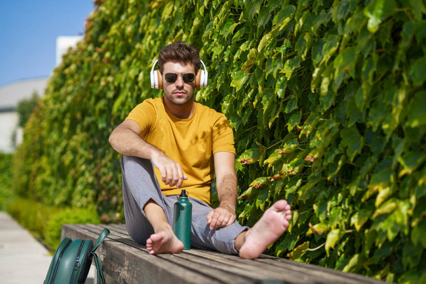 Male sitting outside using an aluminum water bottle, headphones and backpack. - Фото, изображение