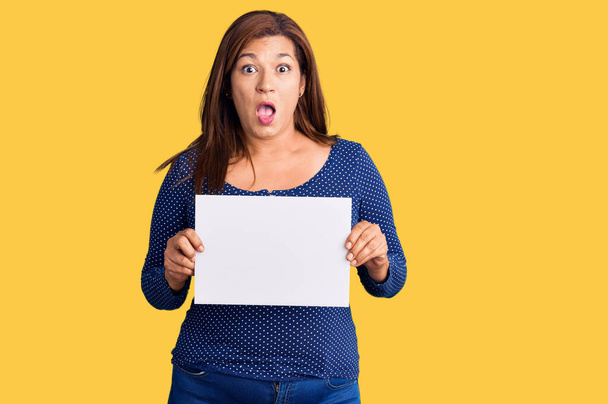 Middle age latin woman holding paper banner with blank space scared and amazed with open mouth for surprise, disbelief face  - Foto, imagen