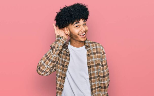 Jonge Afro-Amerikaanse man met Afrikaans haar in casual kleding glimlachend met hand op oor luisterend naar geruchten of roddels. doofheid.  - Foto, afbeelding