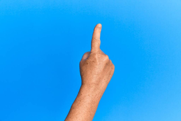 Ręka starszego Hiszpana nad niebieskim odizolowanym tłem licząc numer jeden palcem wskazującym, pokazując ideę i zrozumienie  - Zdjęcie, obraz