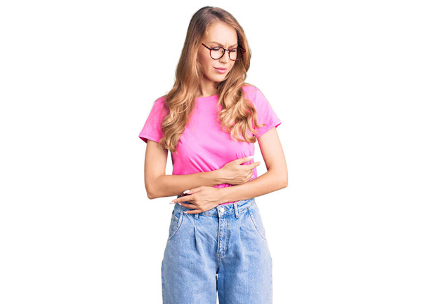 Young beautiful caucasian woman with blond hair wearing casual clothes and glasses with hand on stomach because indigestion, painful illness feeling unwell. ache concept.  - Фото, изображение