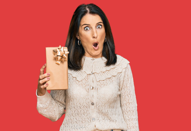Middle age brunette woman holding gift scared and amazed with open mouth for surprise, disbelief face  - Photo, Image