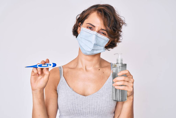 Young hispanic woman wearing medical mask, hand sanitizer gel and thermometer clueless and confused expression. doubt concept.  - Photo, Image