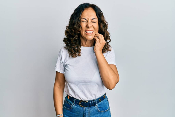 Mujer hispana de mediana edad que usa una camiseta blanca casual tocando la boca con la mano con expresión dolorosa debido a dolor de muelas o enfermedad dental en los dientes. dentista  - Foto, imagen