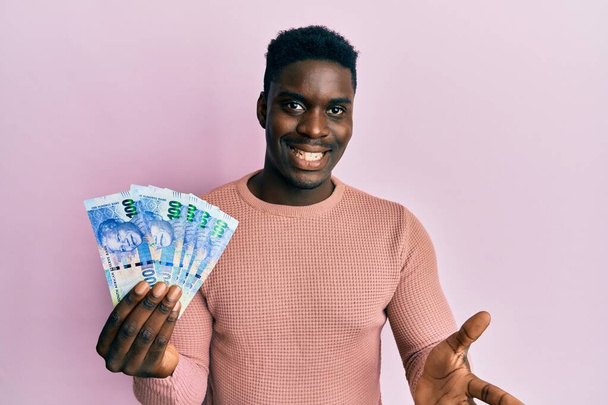 Handsome black man holding south african 100 rand banknotes celebrating achievement with happy smile and winner expression with raised hand  - Fotografie, Obrázek
