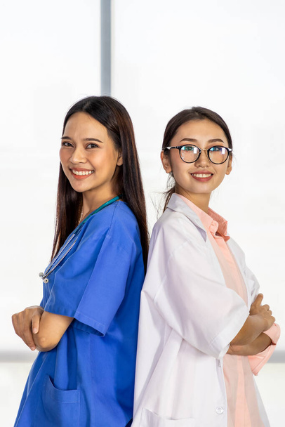 Sırt sırta verip kollarını kavuşturarak gülümseyen ve kameraya bakan iki kendine güvenen Asyalı kadın tıp doktoru. - Fotoğraf, Görsel