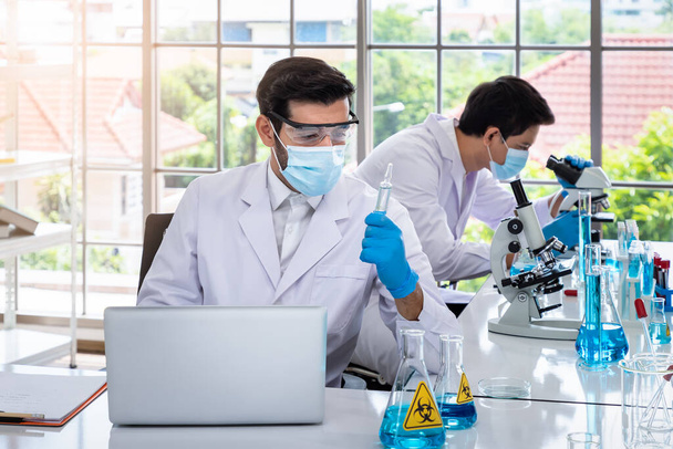 Deux scientifiques masculins asiatiques et arabes travaillant en laboratoire, effectuant des études sur les substances présentant des risques biologiques avec du matériel scientifique et un microscope - Photo, image